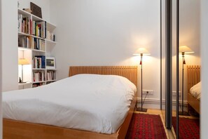 A hotel room with a bed, a bookshelf, a red patterned rug, and a floor lamp.