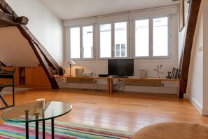 A living room with a TV, wooden beams, large windows, and a colorful rug.