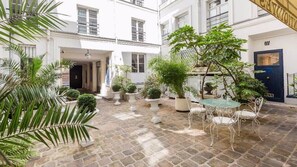 A courtyard with a stone floor, white walls, and potted plants. There is a table with chairs and a door in the background.