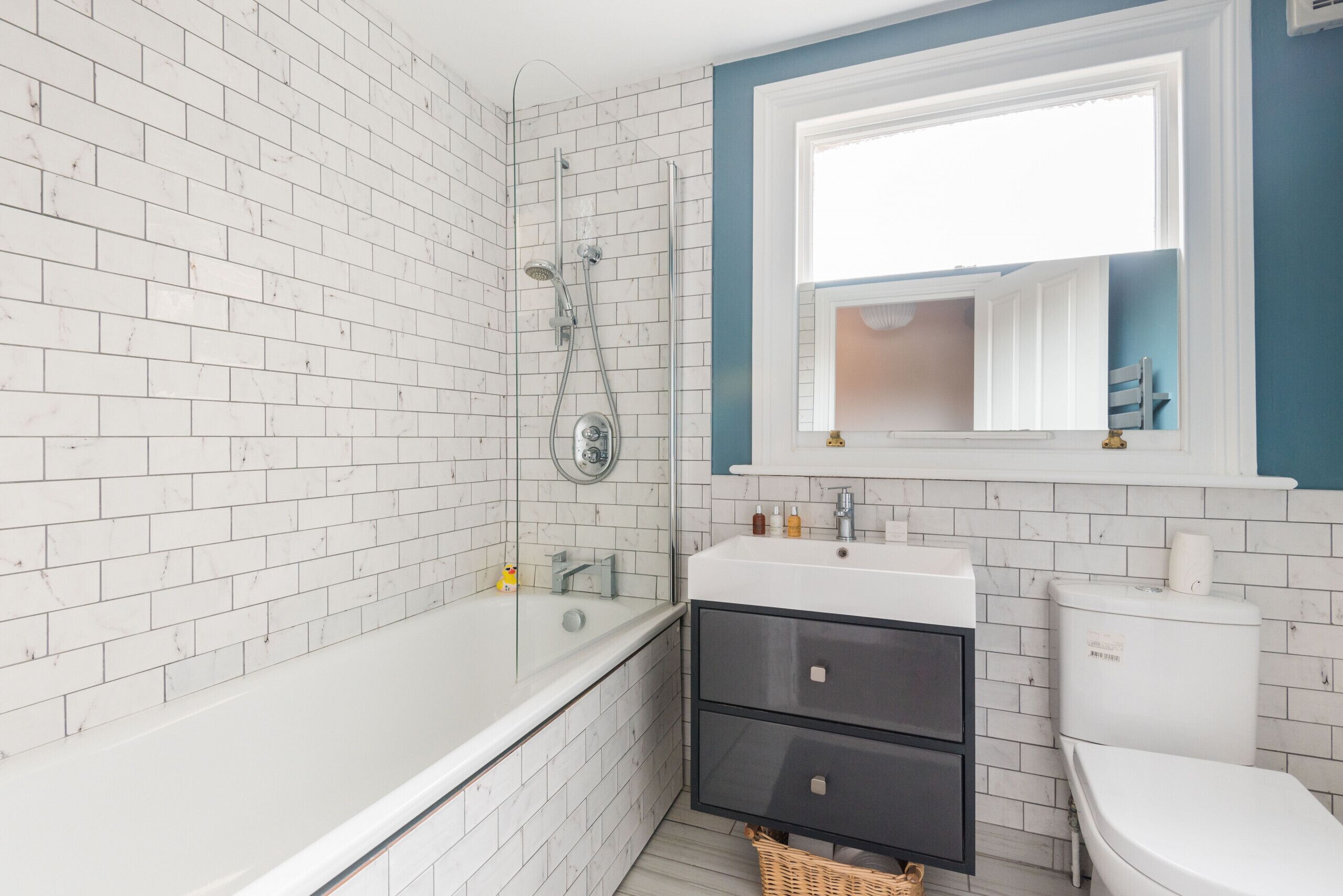 Ein Badezimmer mit weißen Fliesen im U-Bahn-Stil, einer weißen Badewanne, einem schwarzen Waschtisch und einer weißen Toilette. Ein Fenster mit blauem Rahmen.