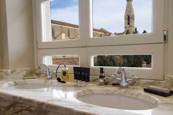 A bathroom with two sinks, a window with a view of a church and tower, and toiletries on the counter.
