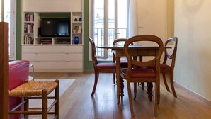A dining area with a wooden table and chairs, a television on a white shelf, and a view of the outside through a glass door. - Coquelicot | Delightful 1 Bedroom Apt near Eiffel Tower (Paris)