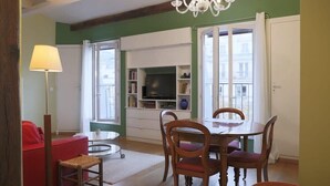 A living room with a red sofa, wooden chairs, a dining table, and a TV on a white entertainment unit. - Coquelicot | Delightful 1 Bedroom Apt near Eiffel Tower (Paris)