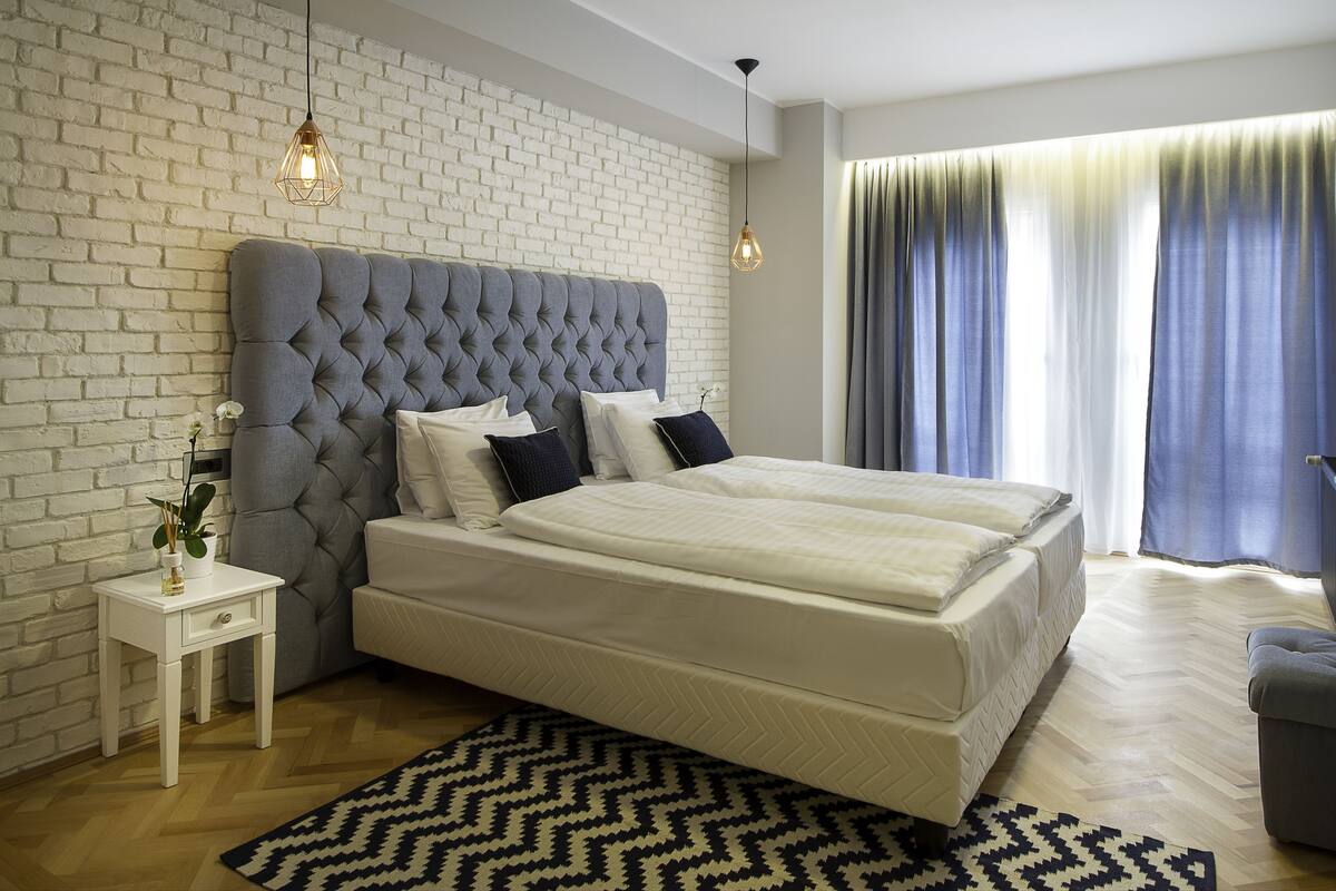 Un dormitorio moderno con un cabecero capitonado, una mesita de noche blanca, una alfombra estampada y un ventanal con cortinas.