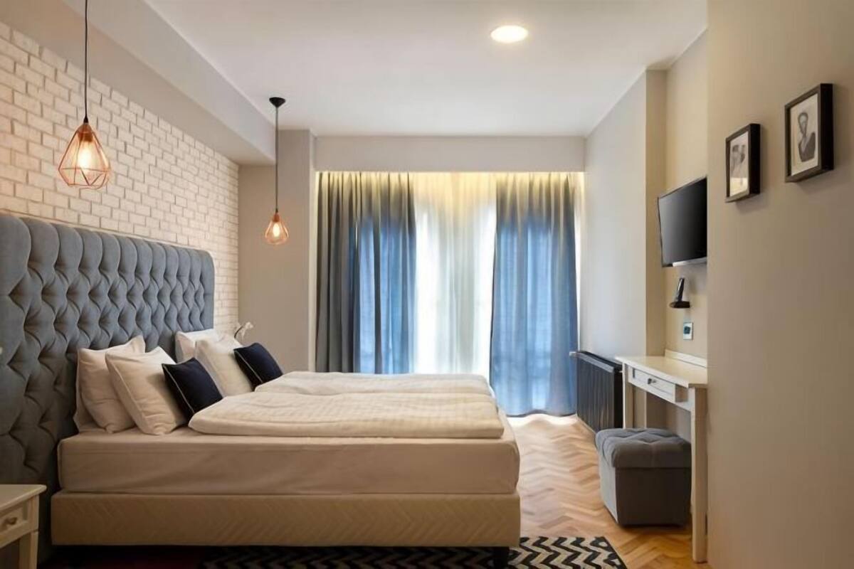 Una cama doble con cabecera capitonada, una mesita de noche, un televisor sobre un mueble bajo y una ventana con cortinas.