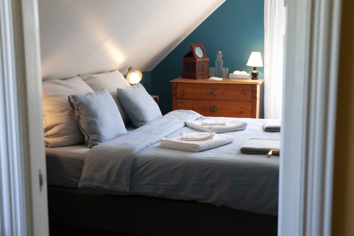 A neatly made bed with pillows and folded towels, a wooden dresser with a lamp, and a wall clock.