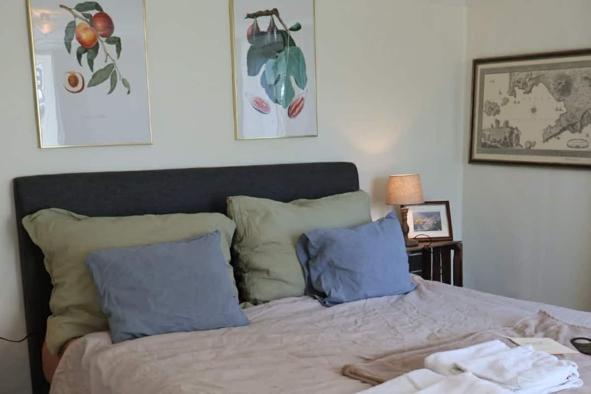 A neatly made bed with a light-colored bedspread and pillows, a framed botanical print on the wall, and a small table with a lamp beside the bed.