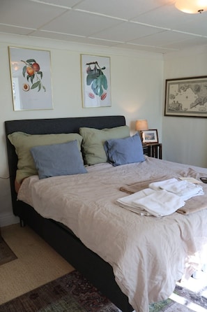 Una cama bien hecha con un edredón de color claro y almohadas, un grabado botánico enmarcado en la pared y una mesita de noche con una lámpara.