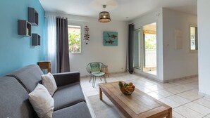 Uma sala de estar moderna com um sofá cinzento, uma mesa de centro de madeira com frutas, uma poltrona verde e uma vista através de uma porta de vidro.