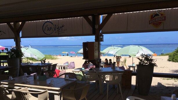 Uma área externa com mesas e cadeiras, com vista para uma praia com guarda-sóis e pessoas.