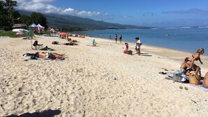Uma praia de areia com pessoas se banhando e tomando sol, barracas e barcos ao fundo.