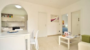 Eine moderne Küche mit weißen Schränken, ein Essbereich mit weißem Tisch und Stühlen sowie eine kleine Sitzecke mit einem grünen Hocker.