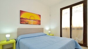 Ein Schlafzimmer mit einem Bett, einem Nachttisch, einer Lampe, einem Sonnenuntergangsbild und einem Fenster mit Vorhängen.