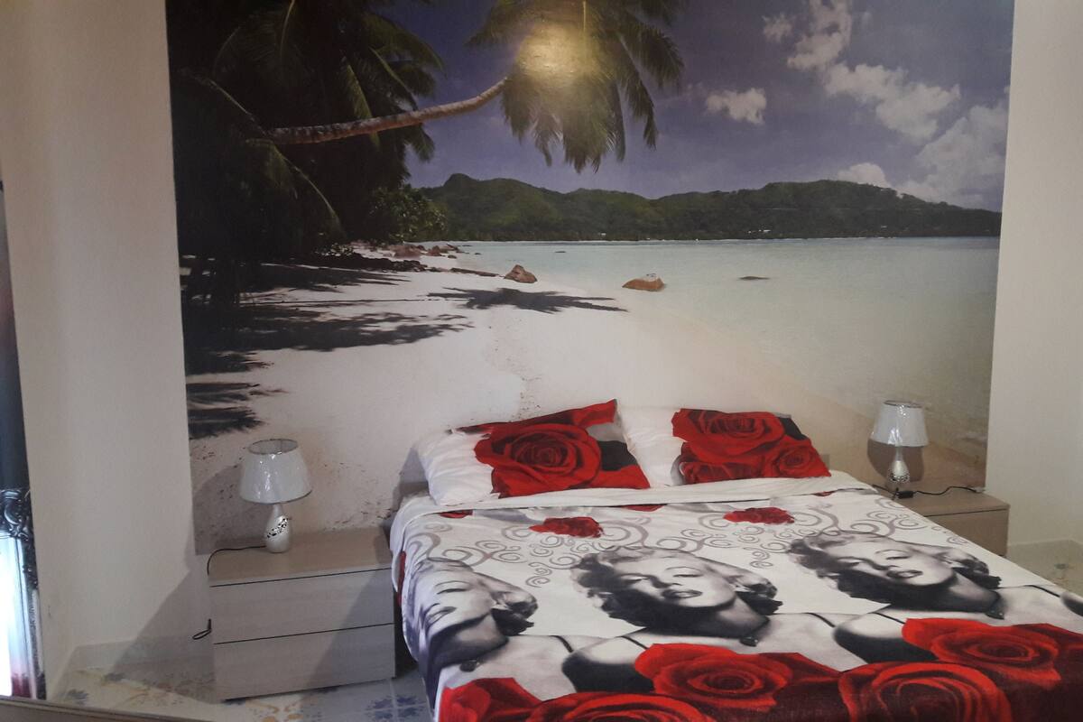 A bedroom with a mural of a beach scene, a bed with rose-patterned bedding, and a nightstand with a lamp.
