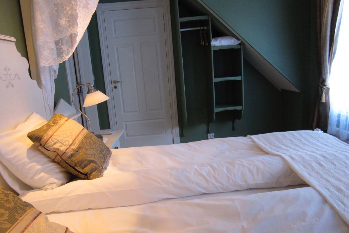 Una cama bien hecha con ropa blanca, un cojín decorativo y una lámpara de noche.