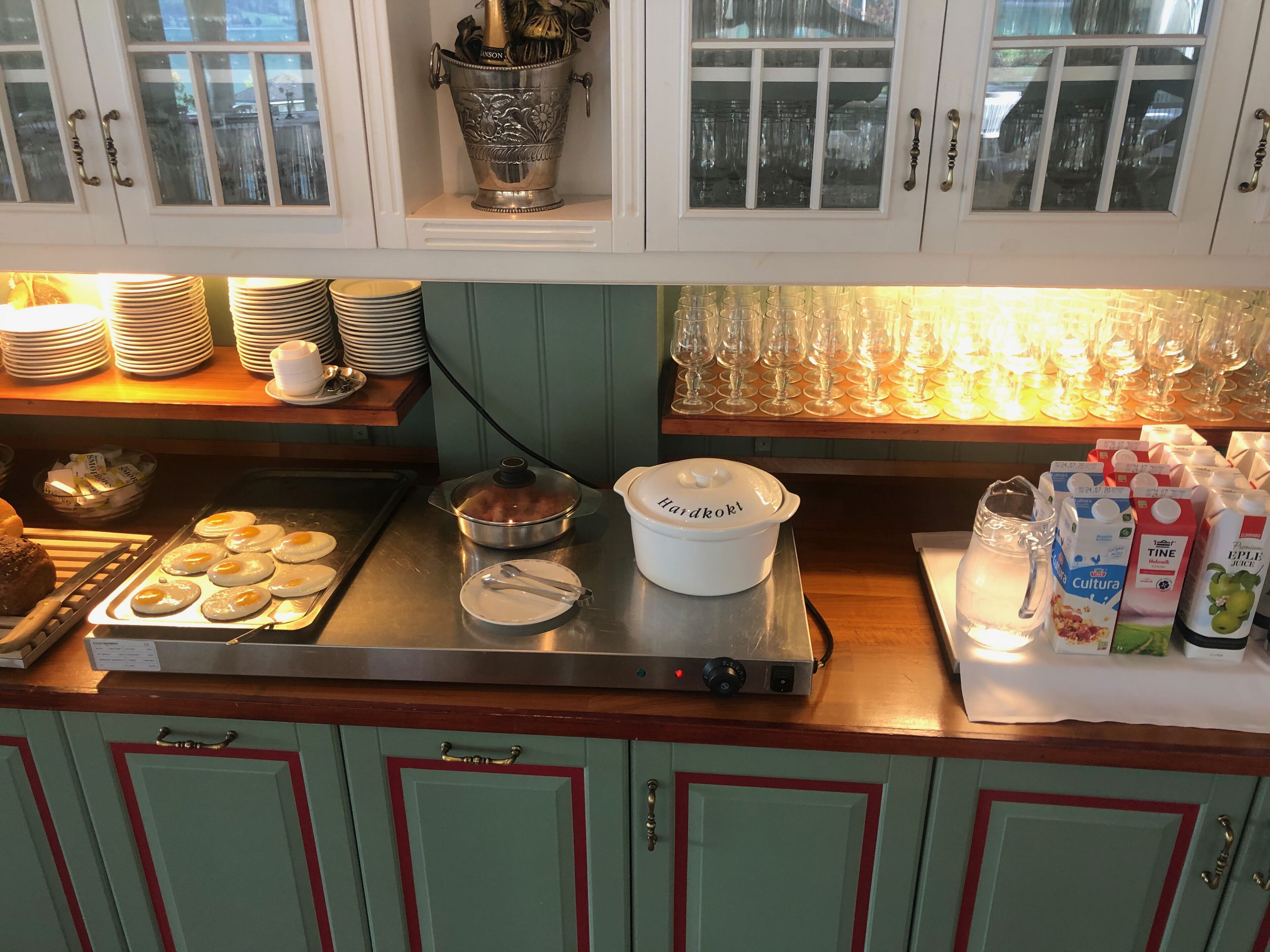 A kitchen with a stainless steel stove, a pot on it, and a white container labeled "Hardkoks". There are stacks of plates and cups on the counter, and a window above the stove.