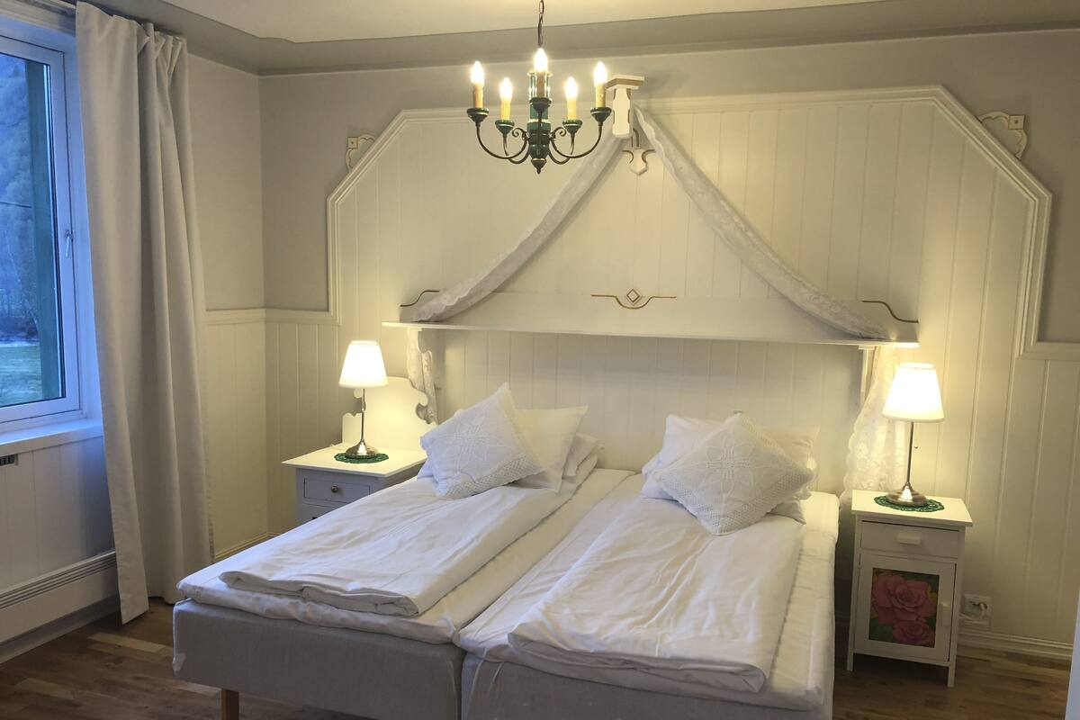 Una cama doble con ropa de cama blanca, dos mesitas de noche con lámparas, una araña y un ventanal con cortinas.