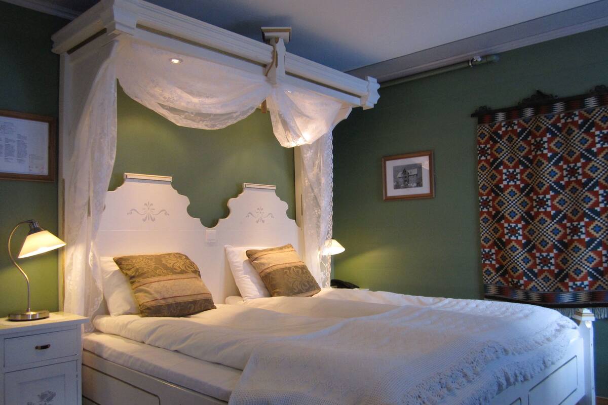 Una cama con dosel y cuatro columnas con ropa de cama blanca y dos almohadas, una mesita de noche con lámpara y una cortina estampada.