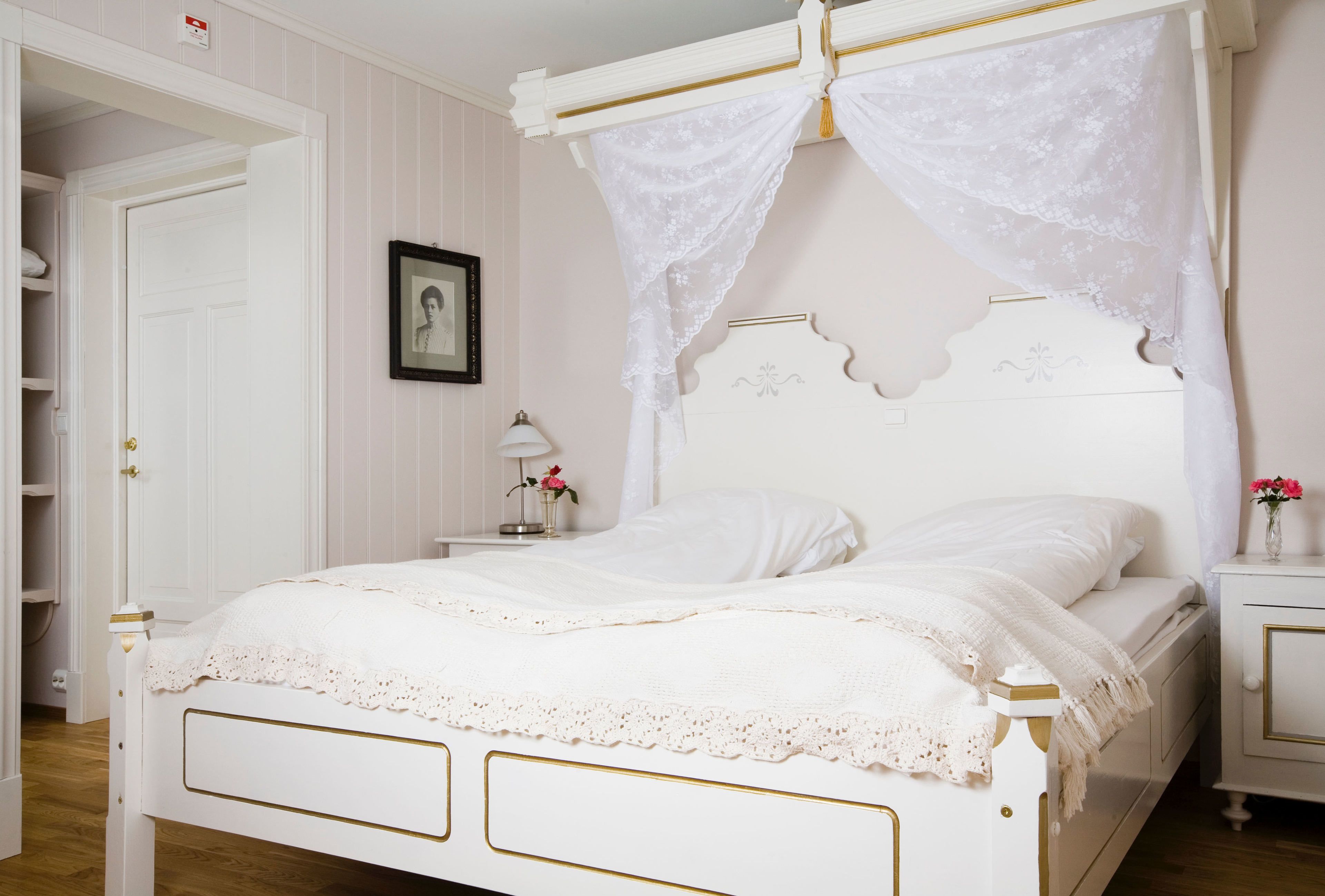 A white four-poster bed with a canopy, a bedside table with a lamp, and a framed picture on the wall.