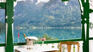 A wooden table with a bottle of champagne, two glasses, and a vase with flowers, set against a scenic mountain lake view. - Hjelle Hotel (Stryn)