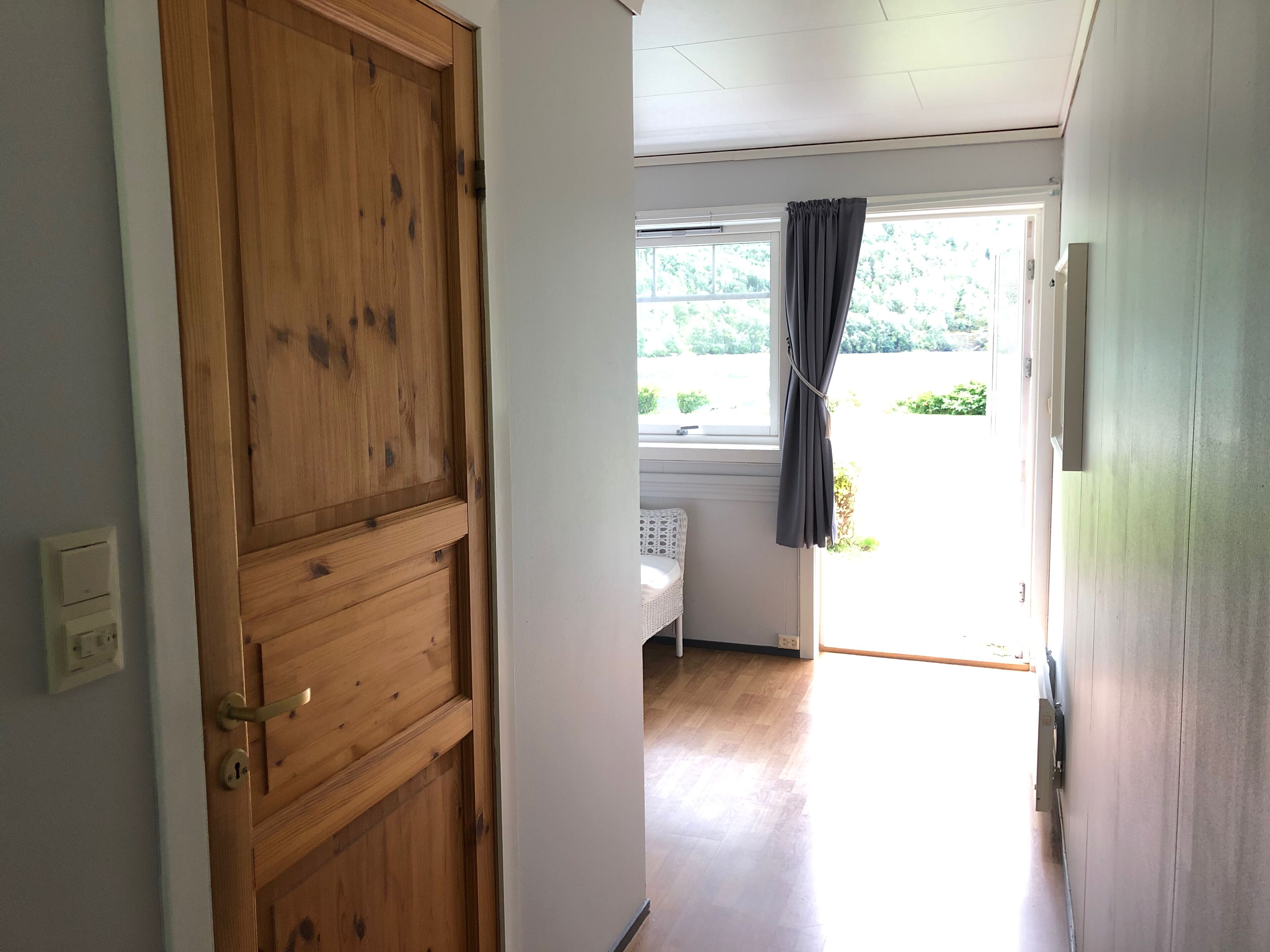 A hallway with a wooden door, a window with curtains, and a bed in the adjacent room.