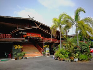 A traditional wooden building with a staircase leading to a balcony, surrounded by potted plants and palm trees. - Pueng Luang Hotel (Damnoen Saduak)