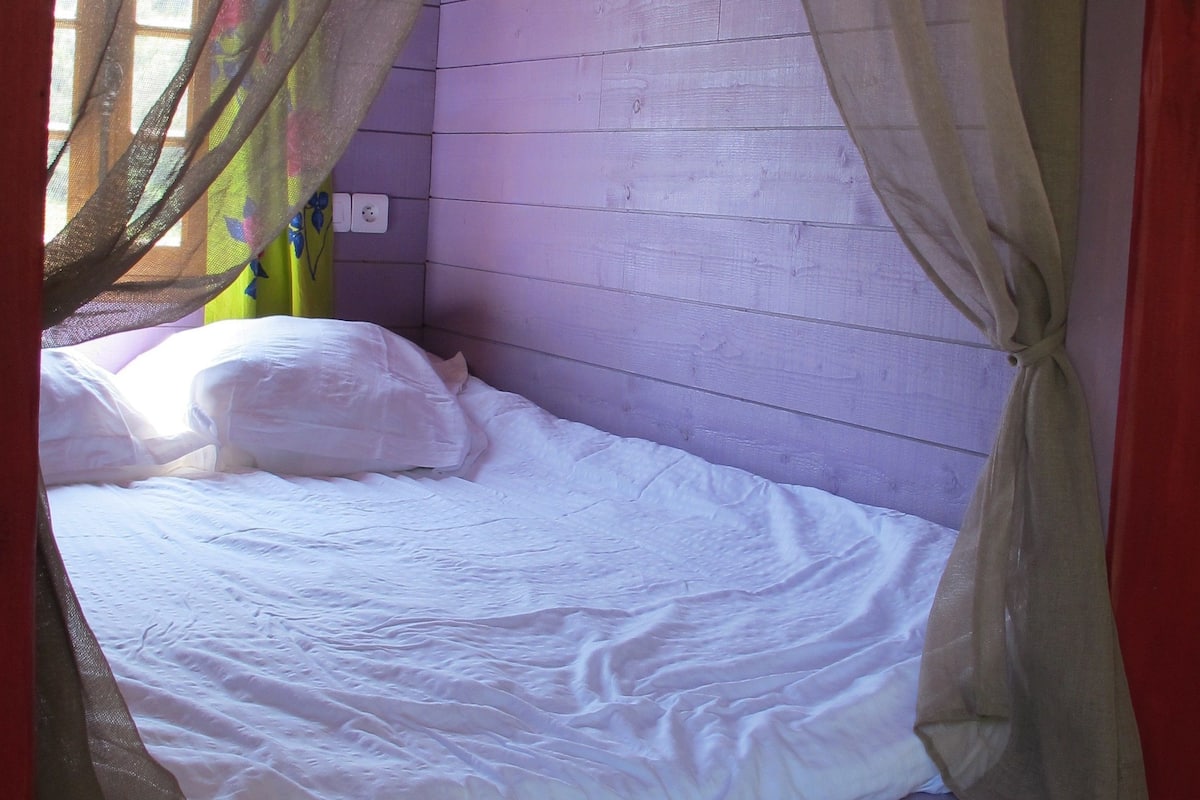 A single bed with a white mattress and pillows, a wooden headboard, and a window with sheer curtains.