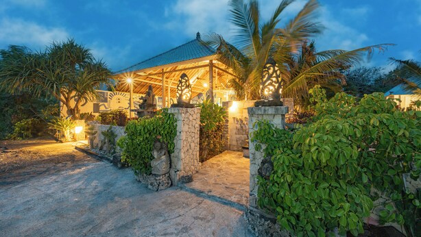 A covered outdoor area with stone pillars, palm trees, and a well-lit building in the background. - The Cubang Huts Lembongan (Lembongan Island)