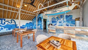 A spacious indoor area with wooden tables and chairs, a ceiling fan, and a mural with nautical themes. - The Cubang Huts Lembongan (Lembongan Island)