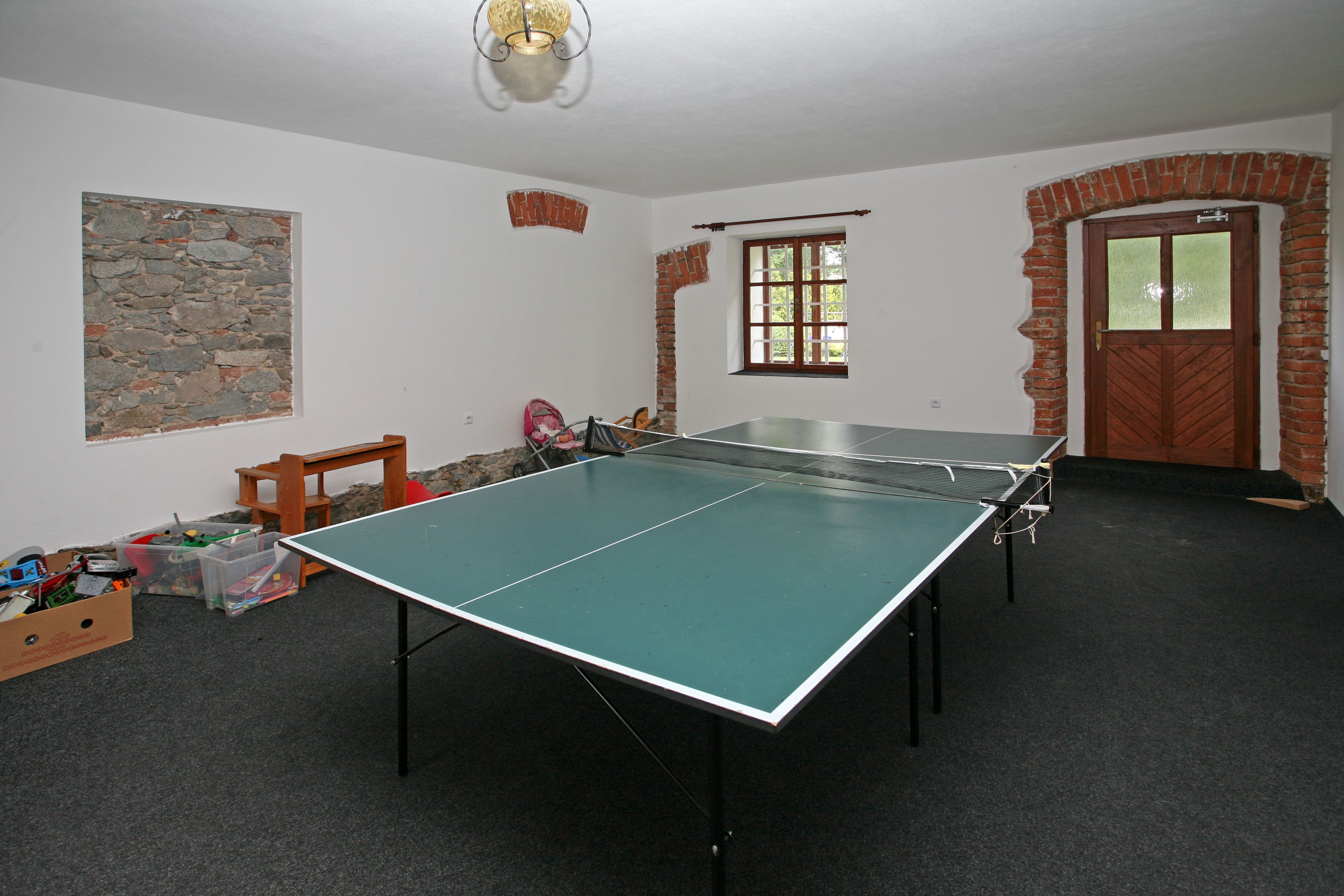 A room with a ping pong table, a stone wall, a wooden door, and a window with a view.