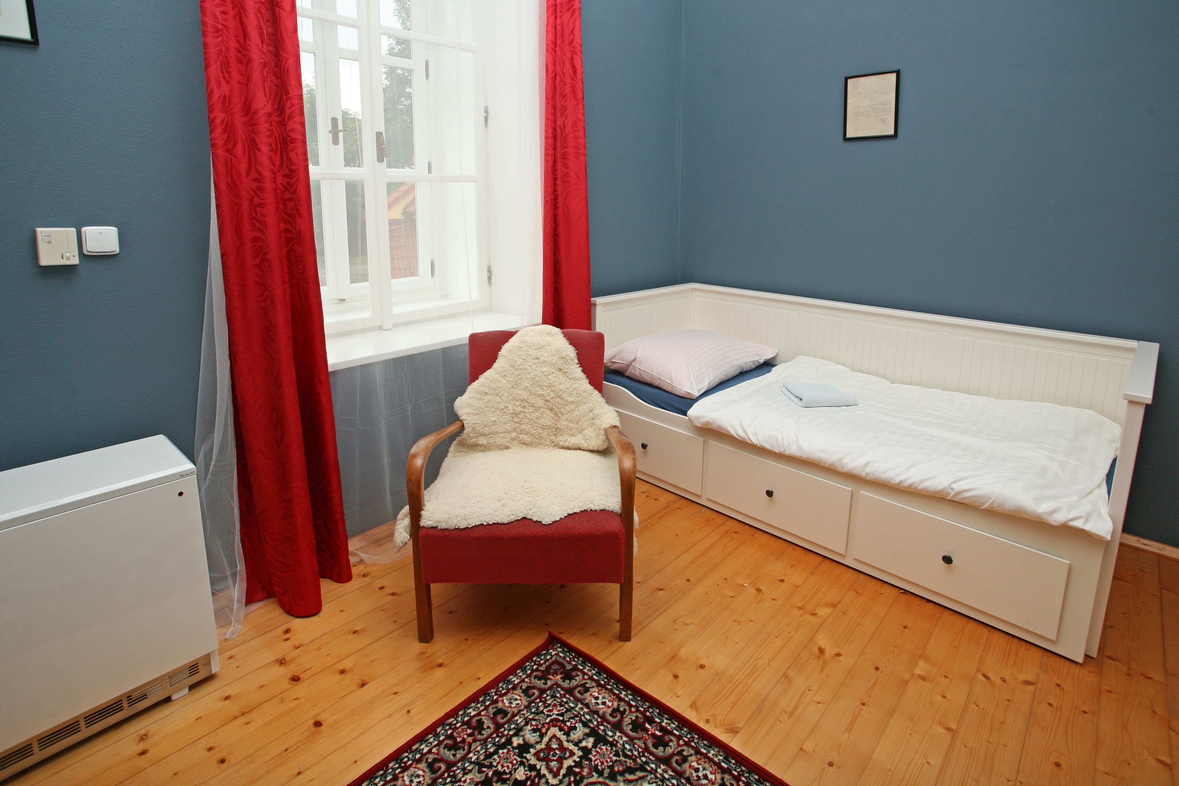 A bedroom with a single bed, a chair with a fur throw, a red and white curtain, a radiator, and a window with a view.