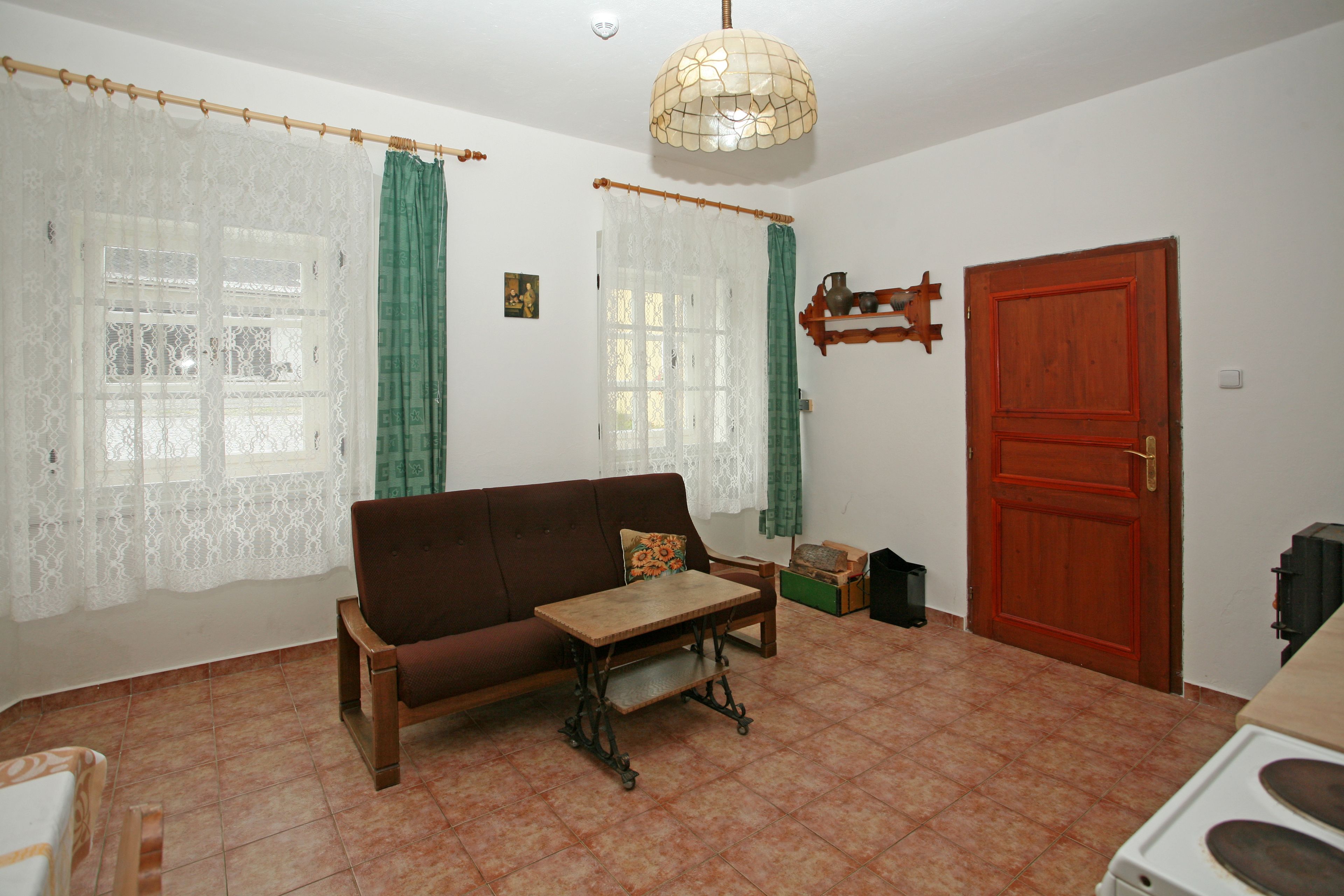 A living room with a brown sofa, a wooden coffee table, a green curtain, a white lace curtain, a red door, and a hanging light fixture.