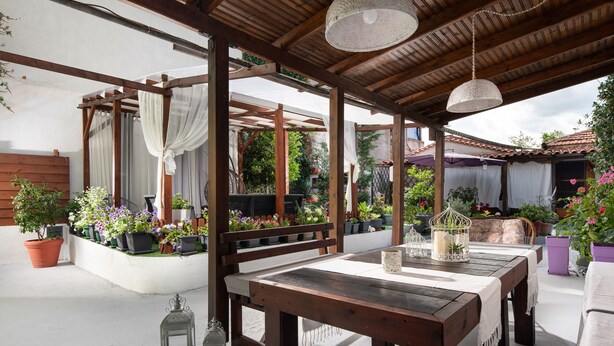 Une terrasse extérieure avec une table et des chaises en bois, entourée de plantes en pot et agrémentée d’une pergola avec des rideaux blancs.