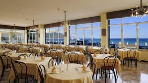 Un comedor con mesas dispuestas para invitados, amplias ventanas que ofrecen vista al mar y una araña colgando del techo.