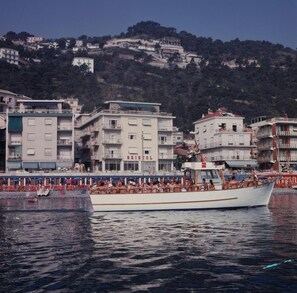 A boat with people on board, docked near a waterfront with buildings and a hillside in the background. - Hotel Bristol (Laigueglia)
