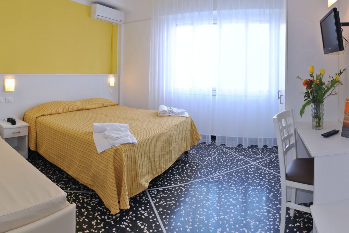 Una camera d'albergo con un letto, una scrivania, una sedia, una TV e una finestra con tende.