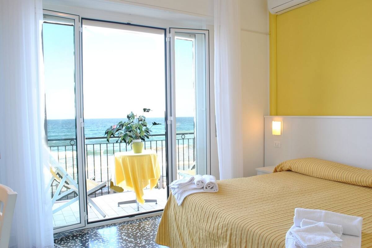 Una camera d'albergo con un letto, un balcone con tavolo e sedie, e vista sul mare.