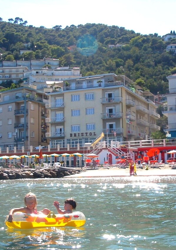 Un hôtel situé sur la côte, avec plusieurs étages, des balcons et un espace de plage équipé de parasols.