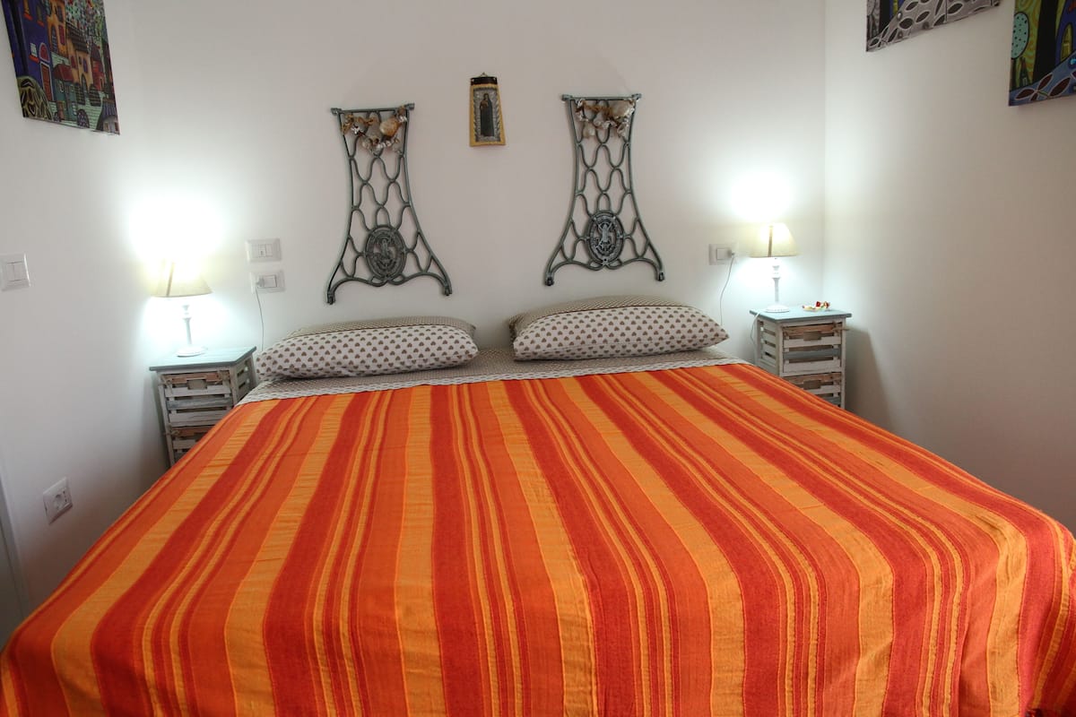 Uma cama com colcha listrada laranja e vermelha, duas almofadas brancas com bolinhas e dois objetos decorativos de metal fixados na parede.