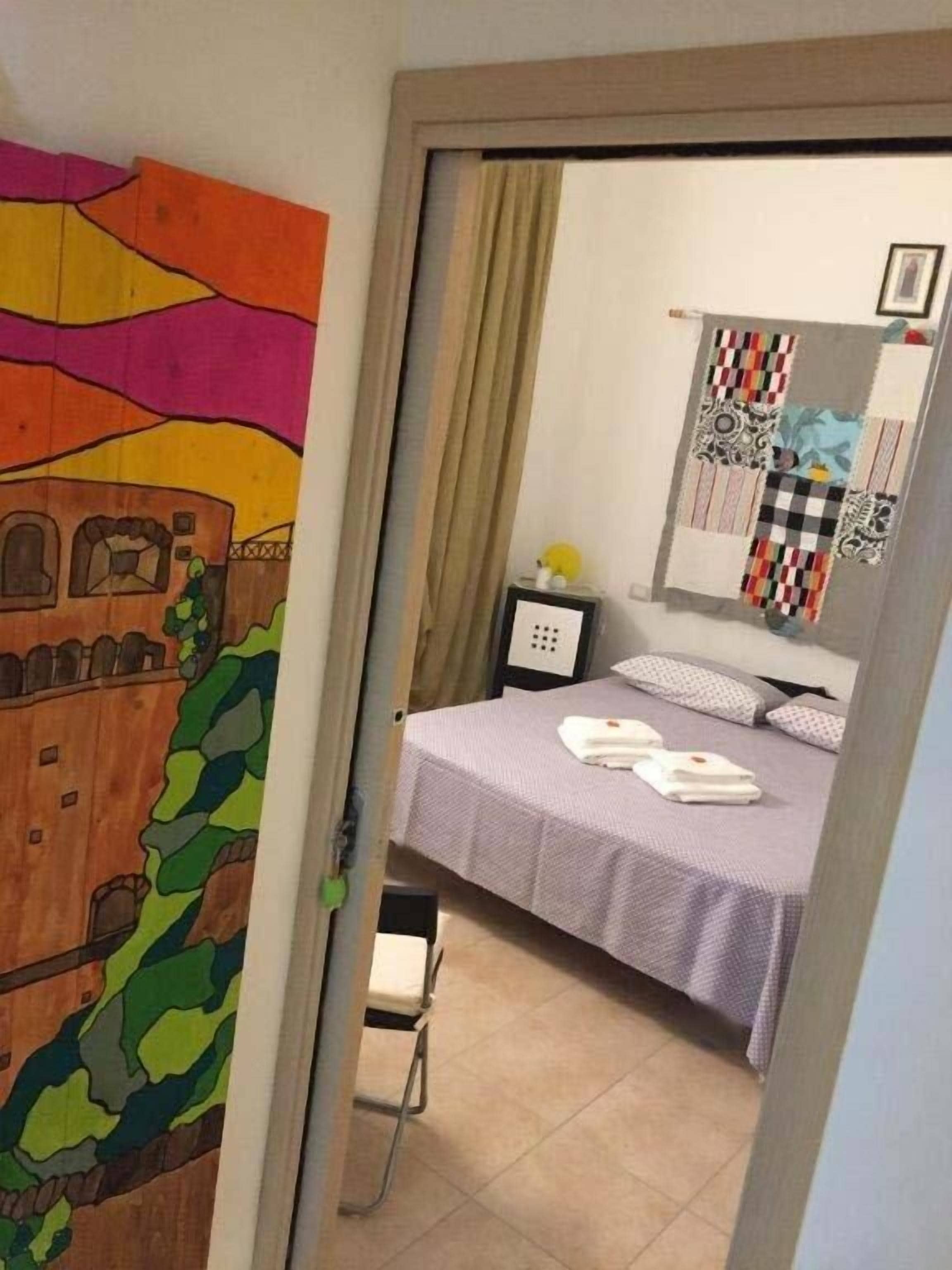 Een slaapkamer met een bed, een nachtkastje, een stoel en een muur met een kleurrijk schilderij.