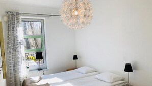 A bedroom with two beds, a window with curtains, a hanging light fixture, and bedside tables with lamps.