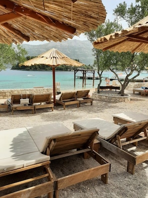 A beachside lounge area with wooden loungers and thatched umbrellas, overlooking a calm sea and distant mountains.