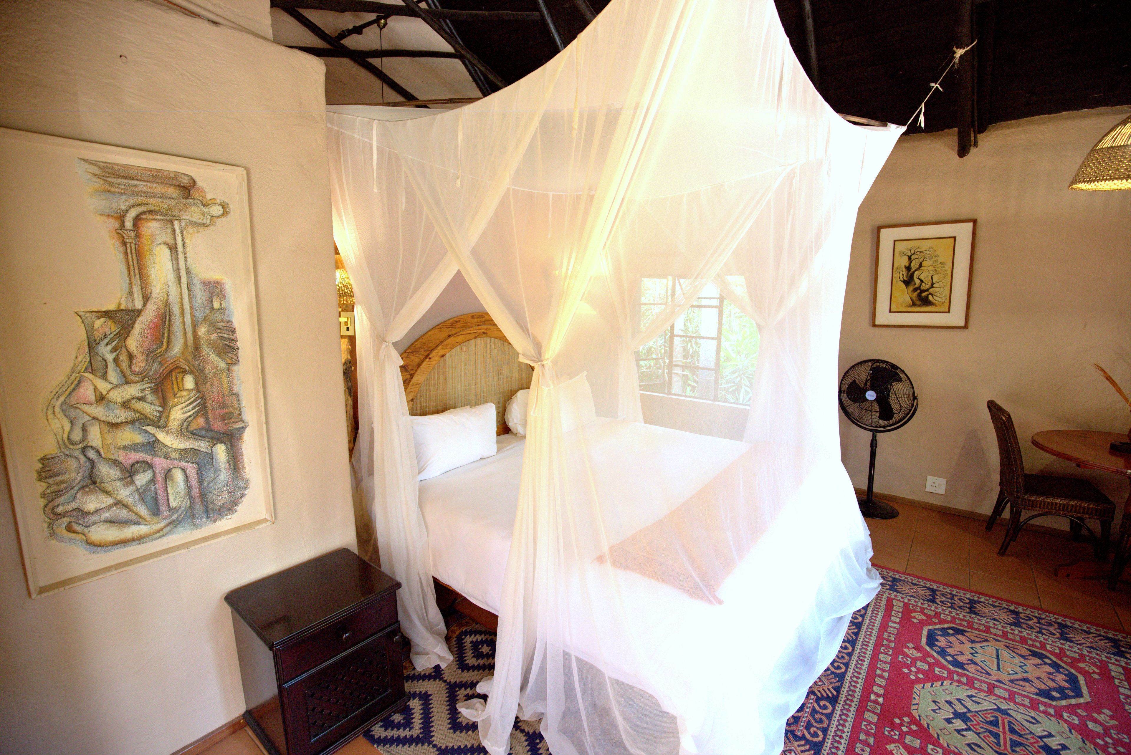 A bedroom with a bed under a white canopy, a framed abstract painting on the wall, a fan, and a patterned rug.