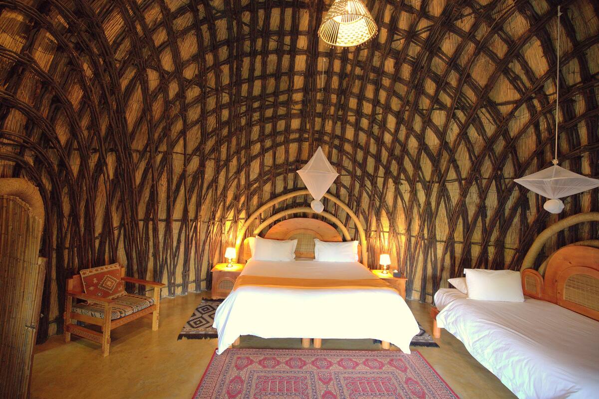 A room with a thatched roof, a bed with white linens, a wooden chair, and two bedside lamps.