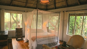 A room with a four-poster bed, wooden flooring, a wicker chair, and large windows with a view of greenery. - Phophonyane Falls Ecolodge (Piggs Peak)
