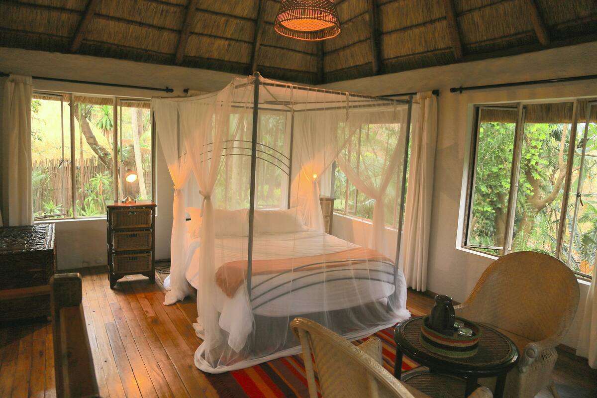 A room with a four-poster bed, wooden flooring, a wicker chair, and large windows with a view of greenery.