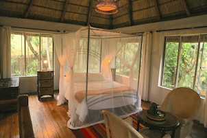 Una habitación con cama con dosel, suelo de madera, una silla de mimbre y ventanas amplias con vistas a la vegetación.