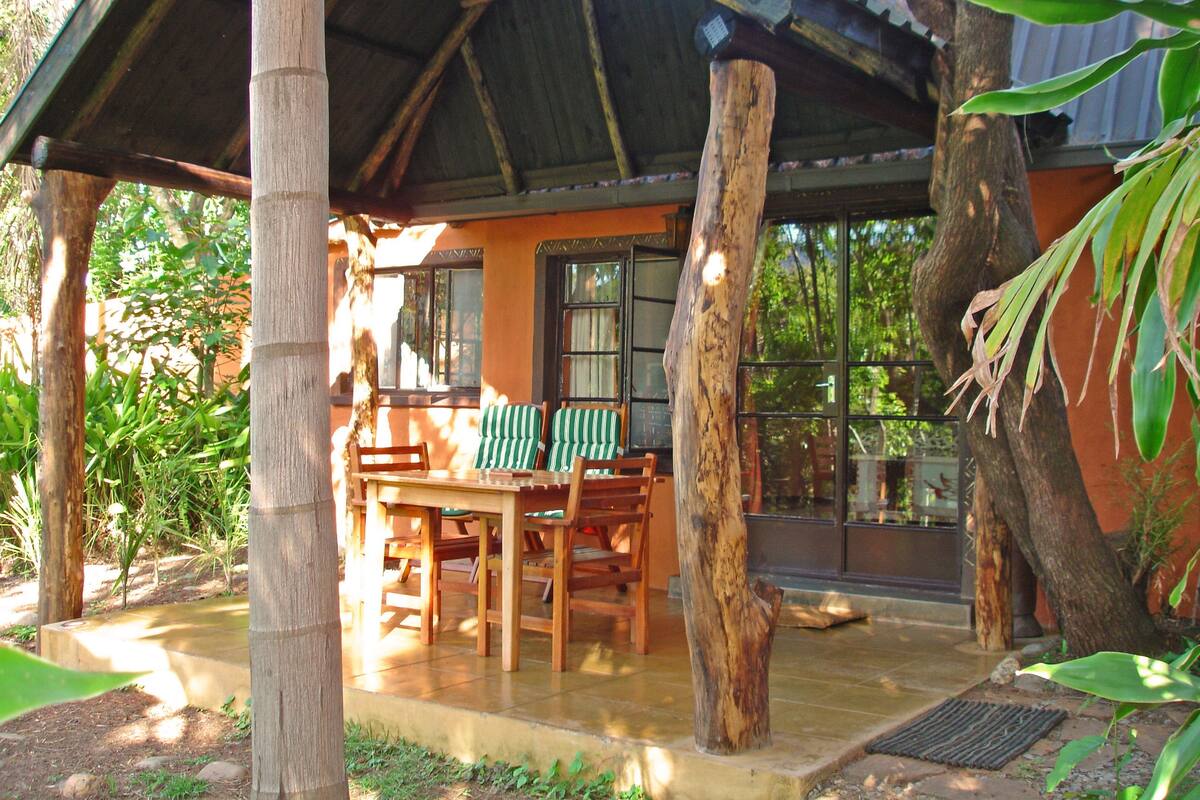 A wooden structure with a table and chairs under a roof, surrounded by trees and a building with large windows.
