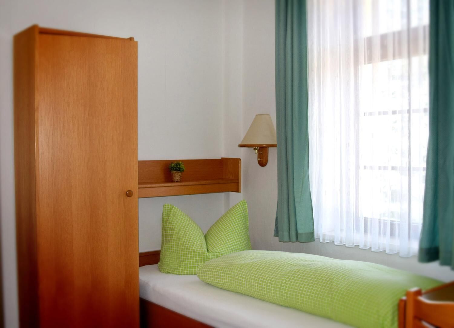 Une petite chambre avec un lit simple, une armoire en bois, une petite étagère avec une plante, une lampe de chevet et une fenêtre avec des rideaux.