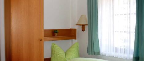 Une petite chambre avec un lit simple, une armoire en bois, une petite étagère avec une plante, une lampe de chevet et une fenêtre avec des rideaux.